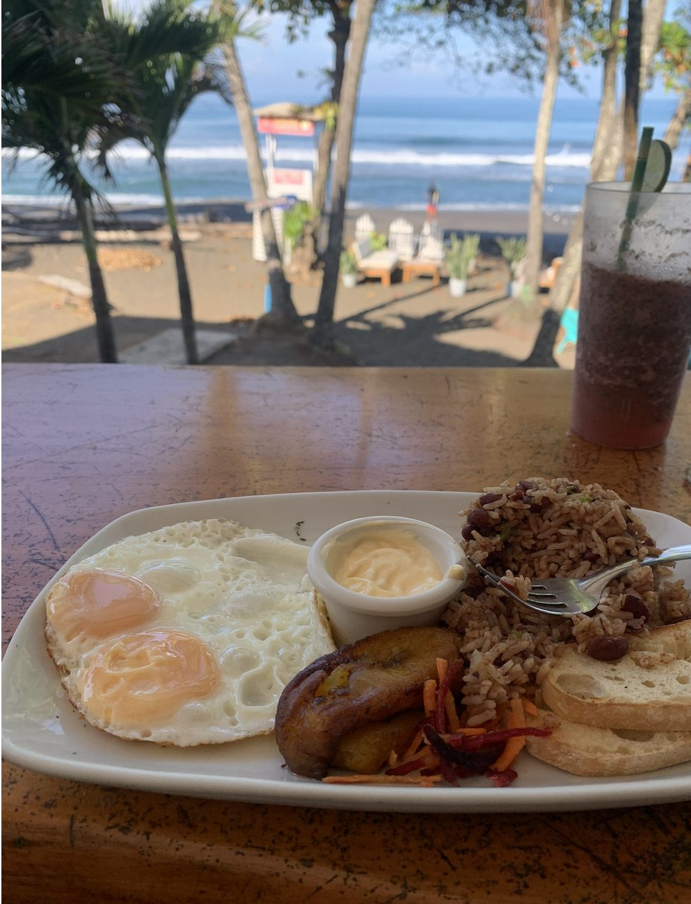 Desayuno típico costarricense con huevos, gallo pinto, plátano maduro y pan, con vista a una playa de arena negra en el Pacífico