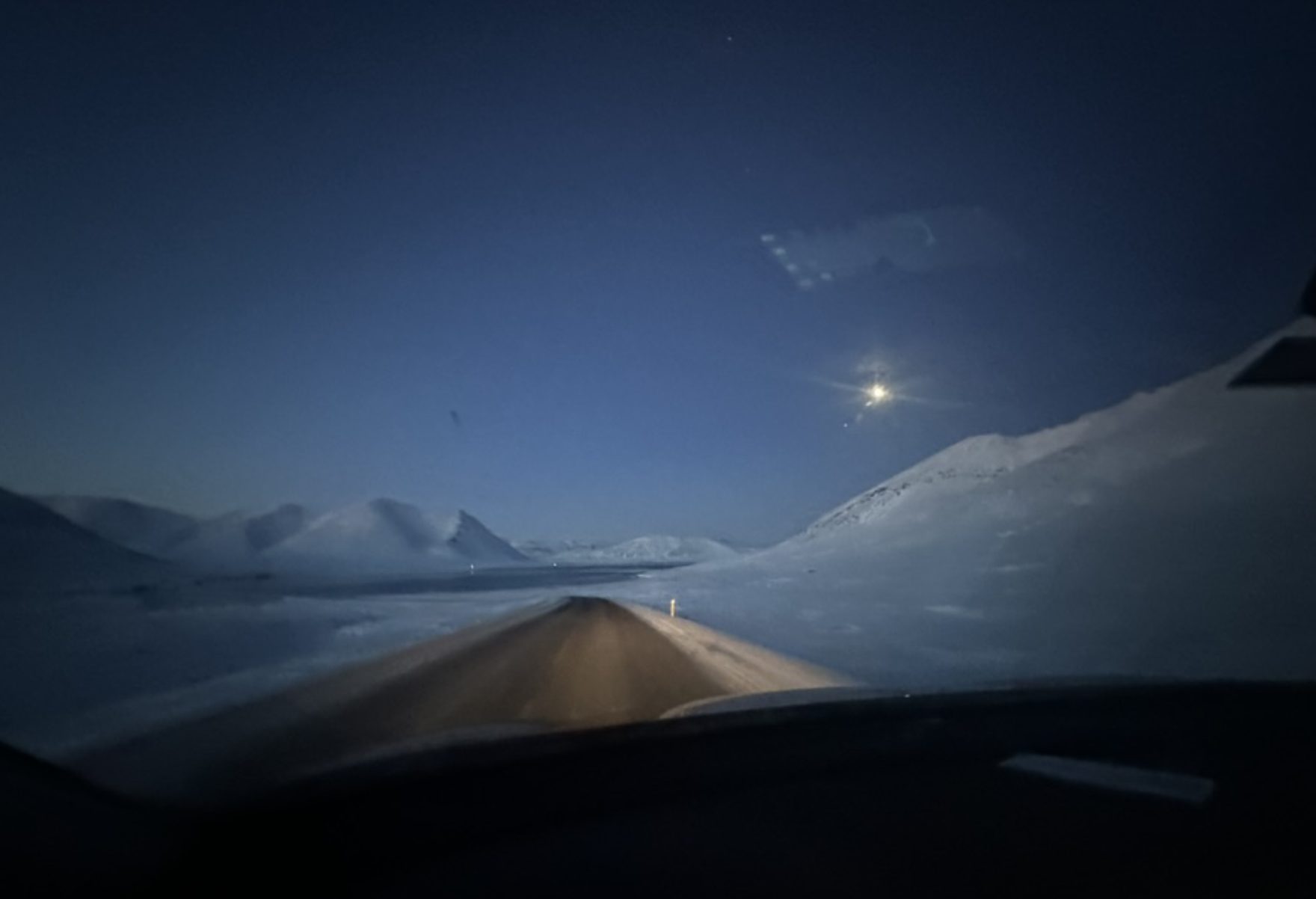 Carretera nevada de noche iluminada por las luces del carro, con la luna en el cielo despejado, Islandia